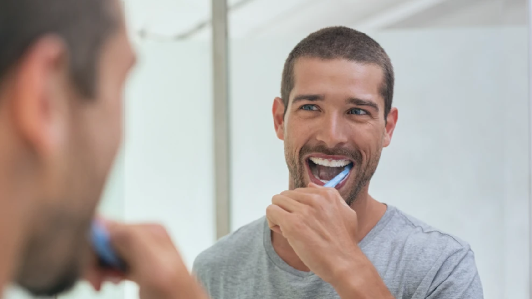Hombre joven cepillándose los dientes frente al espejo con una sonrisa.
